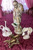 Photo showing all four figurines together on a red floral tablecloth: ceramic servant boy, Capodimonte swans, brass horses, Angel of Compassion figurine.