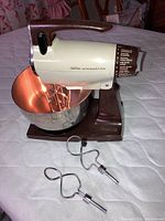 Side angle view of the Sunbeam Mixmaster mixer mounted on its brown base with stainless steel bowl and extra beaters in front, showing the mixer's handle and control switch.