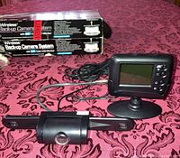 Photo of the wireless backup camera mounted on a license plate bracket next to the 2.5" color LCD monitor with buttons and stand, all on a fabric surface.