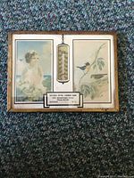 Full front view showing entire wooden framed thermometer with left and right lithograph panels and central thermometer with business plaque.