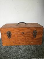 Closed antique wooden tool box showing the front with two metal latches and carrying handle on top.