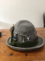 Front and side view of grey wool felt hat with decorative pins, braided green trim around brim, and feathers.