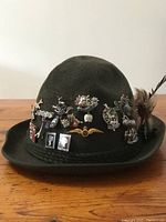 Front view of dark green wool felt hat decorated with various Scottish pins including clan crests and symbols, with detailed image of braided band.