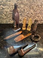 All seven items arranged on dark reflective surface against stone wall background, showing tall man figure, old man and woman figures, doll, bracelet, two pipes, canoe, and letter opener.