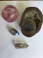 Top view of four items on white background: large brown geode with crystal interior, smaller quartz points, and clear pink swirled glass orb