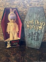 Living Dead Dolls Eggzorcist 13th Anniversary doll in yellow bunny outfit inside original coffin-shaped box against a brick background.