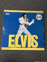 Front cover of Elvis Presley double feature LP album showing Elvis in white jumpsuit with blue background and large yellow 'ELVIS' text. Sticker indicates 2 big LPs priced at 5.98. Visible wear and edge damage on cover.