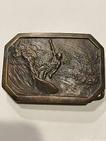 Front view of heavy brass belt buckle with relief of surfer on wave.