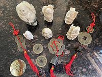 Six ivory-like carved Oriental figurines of various sizes posed on a dark surface, alongside several sets of decorative Oriental coins tied with red tassels and a small shell-like carved piece.