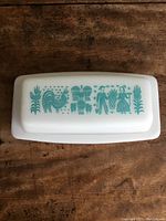 Top view of Pyrex opaque glass butter dish lid showing turquoise folk art design featuring a rooster, plants, and people.