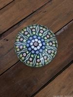 Top view of the round glass paperweight showing multicolored millefiori floral patterns with a green background.