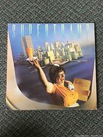 Front cover of Supertramp Breakfast In America vinyl LP album showing waitress with orange juice glass and cityscape background.