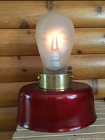 Front view of frosted glass mannequin head lamp lit internally showing facial features and head texture on a red base with brass collar.