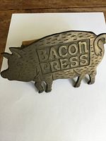 Front view of pig-shaped brass bacon press with 'BACON PRESS' embossed on the side.