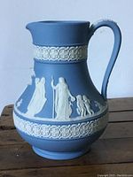 Blue Wedgewood jasperware pottery jug with white classical relief bands and detailed figure decoration, showing profile from side with handle and pouring spout.