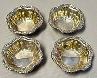 Four small sterling silver salt cellars with detailed repoussé rim, arranged on a table surface, showing all items in the lot.