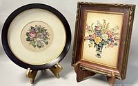 Both floral artworks shown side by side on wooden stands, displaying oval needlepoint and rectangular print framed artworks with minor frame wear