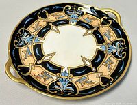 Top down view of round porcelain serving cake plate showing detailed gilded, blue and black painted pattern with two gold handles.