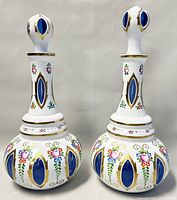 Pair of white-to-blue cut glass Bohemian Czech Art Deco decanters with hand painted floral patterns and gold trim, standing side by side with stoppers in place.