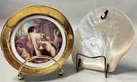 Photo showing miniature decorative plate with cherub painting and gold border, along with a shiny mother-of-pearl shell on display stands