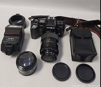 Minolta AF7000 Maxxum 35mm film camera with 100-200mm lens attached, external flash, additional 50mm lens, lens caps, and black leather pouch arranged on a white background.