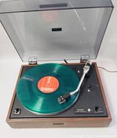 Front angle showing wood veneer base, black top control panel, turntable platter with green vinyl record, and transparent dust cover.