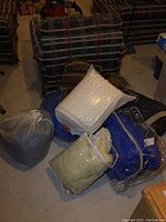 Photo showing assorted sleeping bags, foam toppers, and throw pillows on floor in front of plaid furniture.