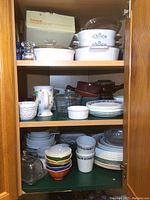 Corner cupboard with multiple shelves containing Corning Ware pieces with blue floral pattern on the top shelf along with Corelle plates and bowls, colorful small bowls stacked and onion soup bowls.