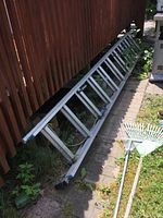 Photo showing the full length of the aluminum extension ladder placed on the ground next to a wooden fence, with garden tools nearby.