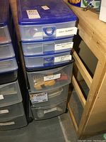 Photo showing two blue plastic 3-drawer storage bins labeled with contents such as saw blades, drill bit/socket set, screwdriver set, and drill bits set. Bins are stacked and positioned beside a wooden frame.