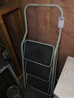 Folded white metal 3-step painter's ladder with black rubber tread on steps, leaning against a wall in a basement setting.