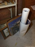 Several framed photos and prints standing against wall with two rolled posters outside frames.