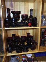 Wide view of multiple ruby red glass items including pitchers, goblets, candlesticks and smaller containers arranged on wood shelves.