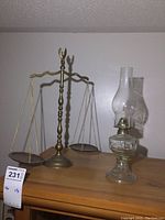 Full view of a brass balance scale and glass oil lamp on a wooden surface against a white wall.