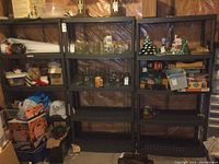 Front view of three black resin shelving units with various household items on them, against a plastic-covered wall in a basement area.