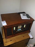Front view of the wooden stereo cabinet with analog radio dial and speakers on both sides, placed on wooden furniture with remote and manual on top.