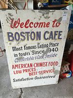 Front face of vintage painted wood sign for Boston Cafe with text showing wear and concrete splatter marks