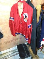 Photo showing full red sports jacket hanging with patches visible on chest and sleeve, with part of a black case on the floor beneath.
