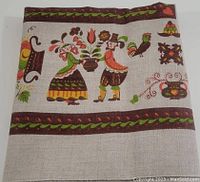 Folded linen tablecloth showing full folklore design of dancing couple, rooster, flowers, and decorative borders against natural linen background.