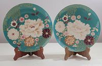 Two fine bone china plates on wooden stands showing full floral pattern on teal background, one plate has slightly different floral arrangement