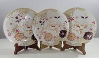Three Maxwell and Williams fine bone china plates displayed on wooden stands, showing full floral and gold accent design.
