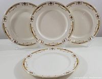 Four white porcelain dinner plates arranged standing and one lying flat, showing full decorative border and gold rim.
