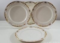 Six round ceramic dinner plates with a decorative gold and reddish-brown border featuring urn and garland motifs, arranged on clear plate stands against white background.