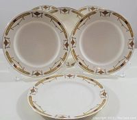 Six white porcelain salad plates with gold, red, and brown decorative border featuring urn motifs arranged on stands.