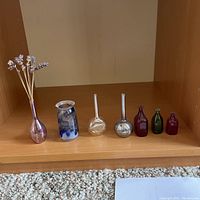 Photo showing 7 miniature coloured glass vases arranged in a row on a wooden shelf.