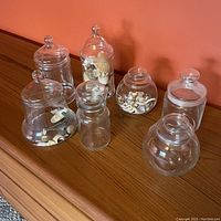 Photo of seven clear glass apothecary jars arranged on wooden surface, some containing assorted natural sea shells
