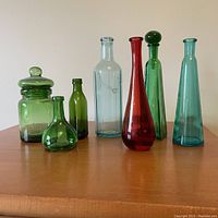 Full set of 7 assorted coloured glass bottles showing various shapes, sizes and colours on wooden surface.