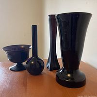 Four black amethyst glass pieces on a wooden surface against a white wall, showing the shapes and relative sizes of three vases and one bowl from a side angle.