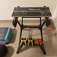 Photo showing the Black & Decker Workmate 225 portable workbench standing on carpet. Two yellow swivel grip clamps attached, and underneath the workbench on the lower base shelf are two swivel grip clamps and a sealed package of four orange swivel grip clamping accessories.