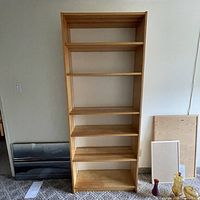 Front view of a tall pine wood bookcase showing 5 shelves, set against a plain wall with some items leaning beside it.